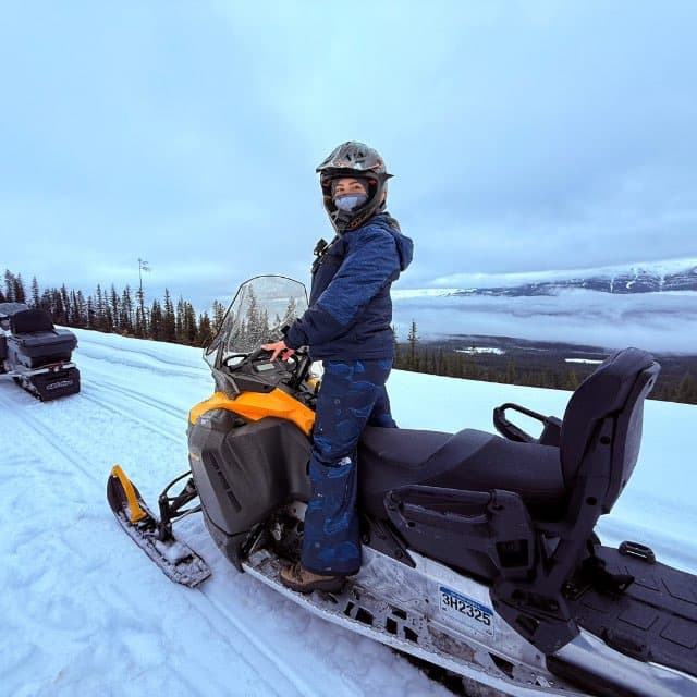 Shaye Davoodi riding a snowmobile in a snowy mountain landscape.